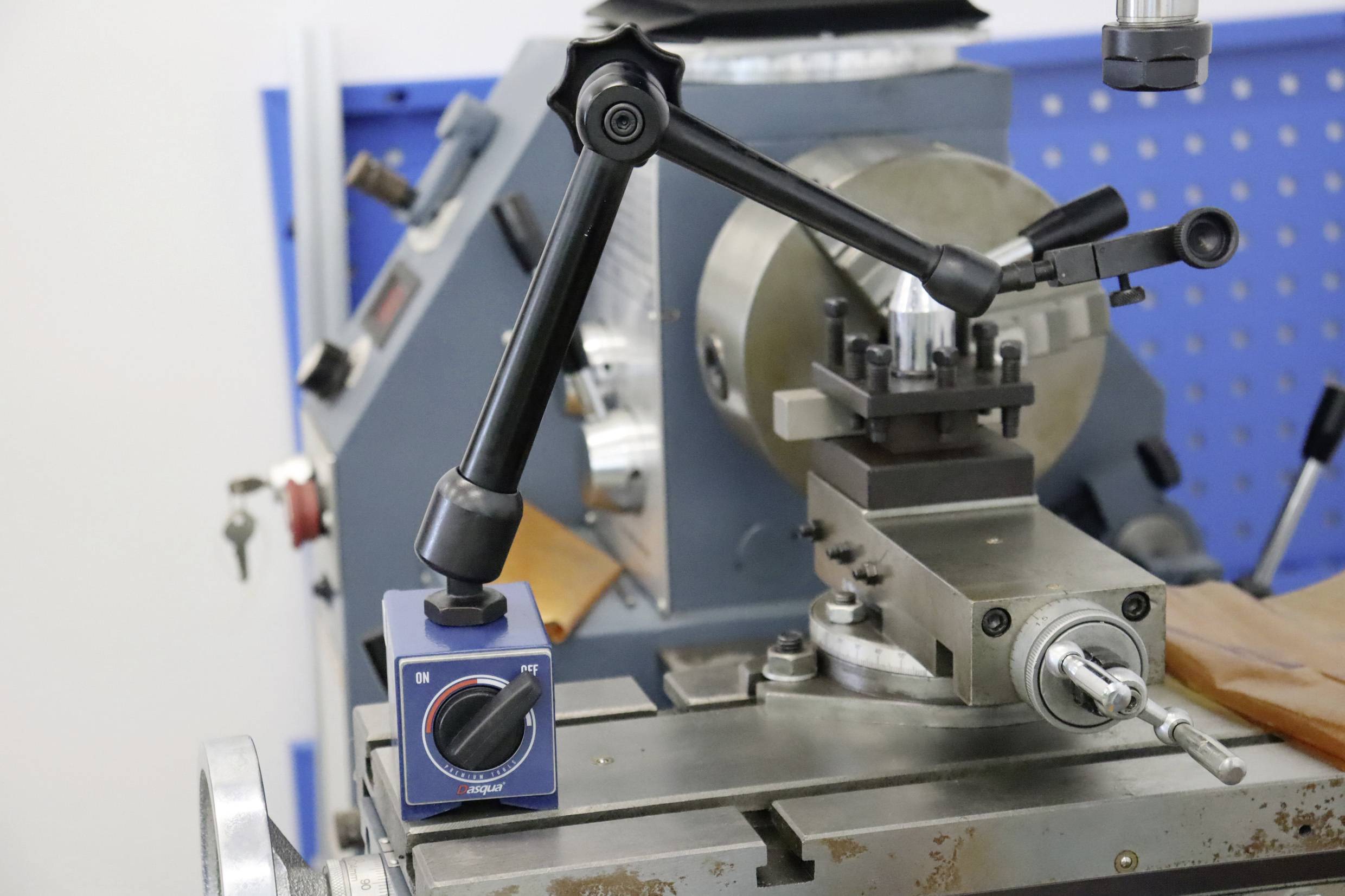 Tokarka z ramieniem montażowym i niebieskim pokrętłem regulacyjnym. W tle widoczna niebieska ściana narzędziowa z wzorem otworów.