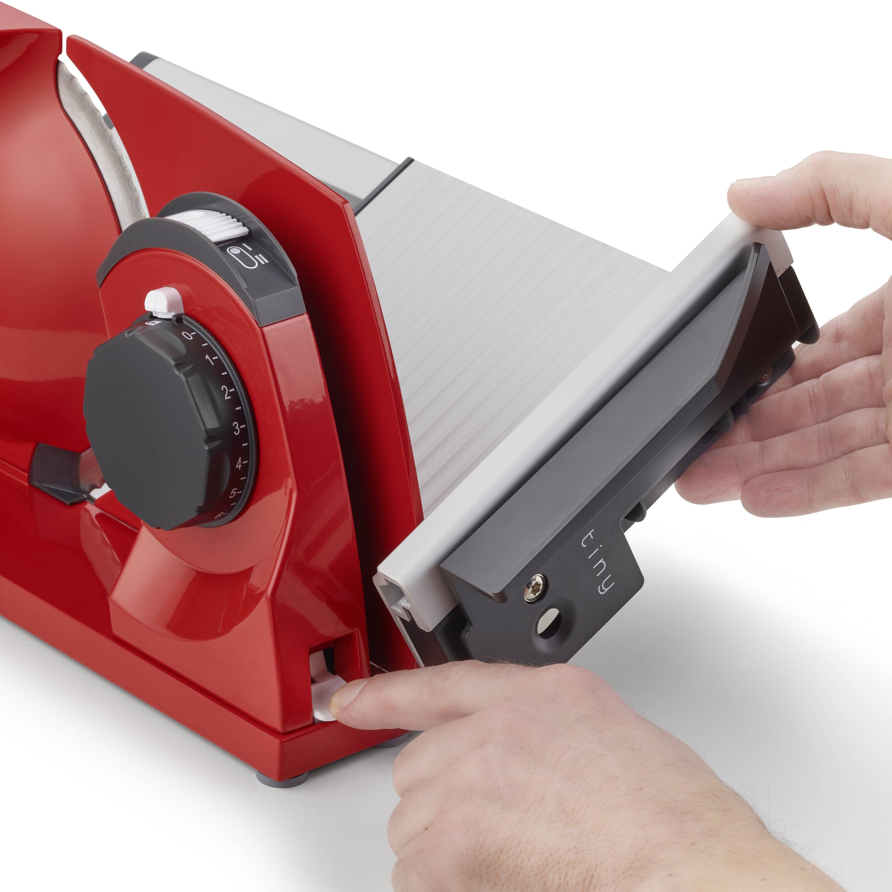 Osoba obsługuje czerwoną wielofunkcyjną krajalnicę z dużą skalą i pokrętłem do regulacji grubości plasterków. Trzyma osłonę noża w stabilnej pozycji.
