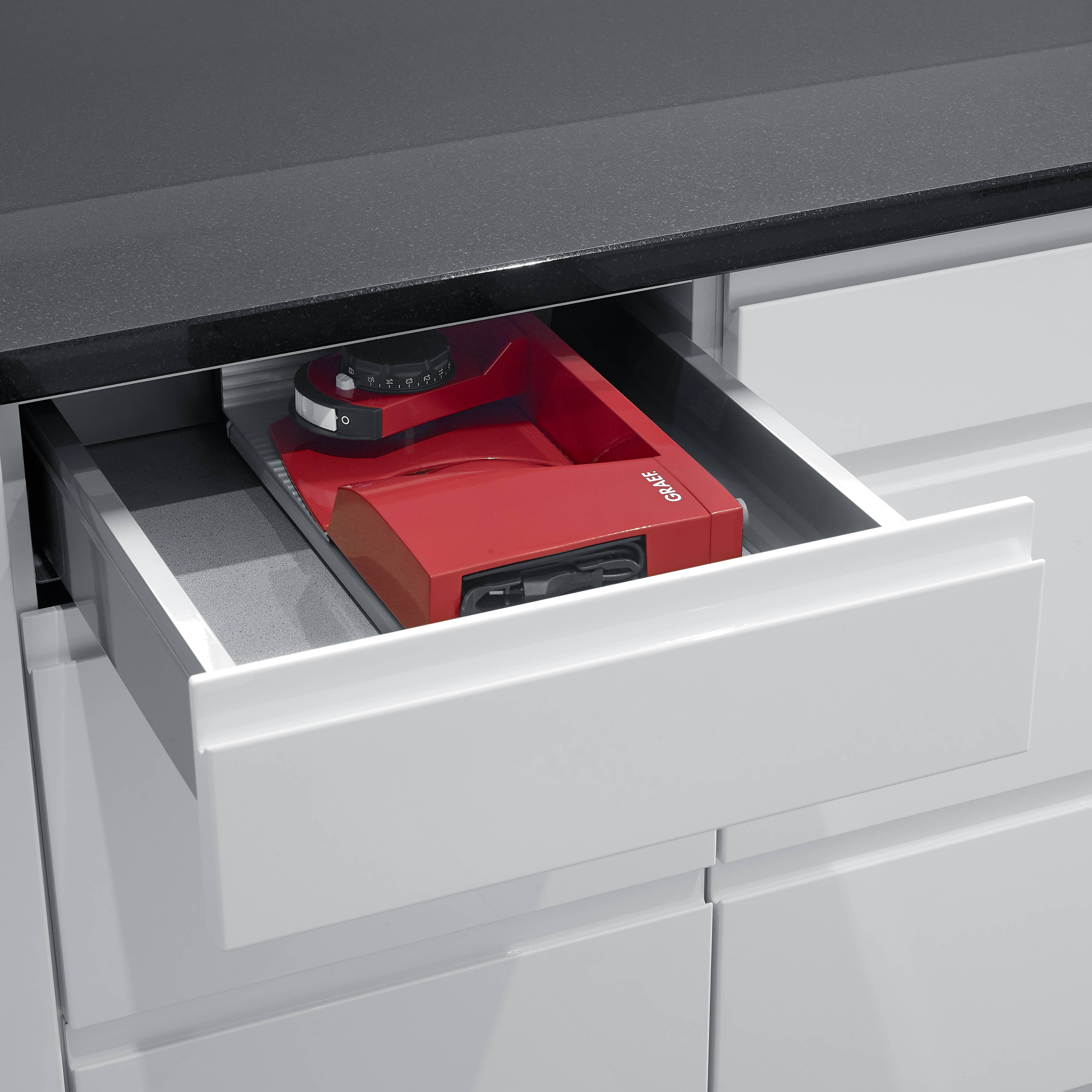 Na otwartej szufladzie kuchennej, pod czarnym blatem, widać czerwoną wagę kuchenną.