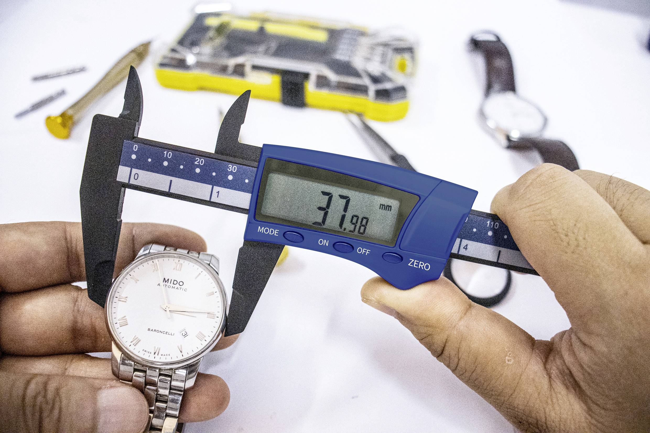 Osoba mierzy średnicę zegarka za pomocą cyfrowego suwmiarki. Wyświetlacz wskazuje 37,98 mm. Na tle leżą narzędzia.