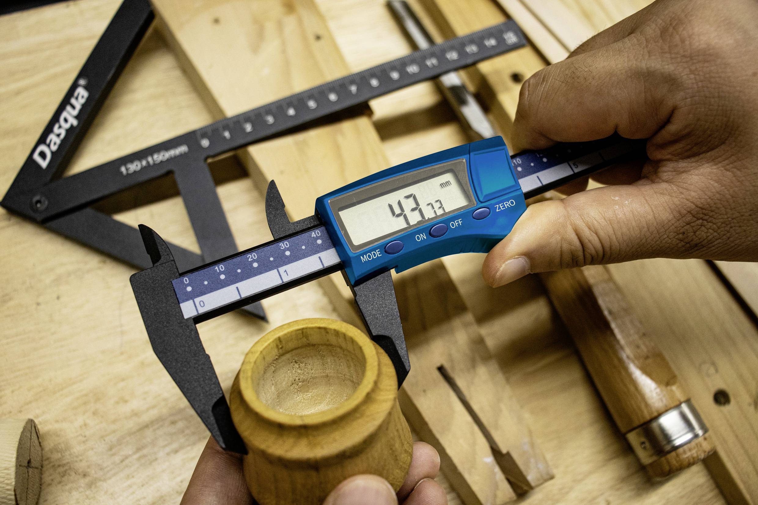 Dwie ręce mierzą średnicę drewna suwmiarką cyfrową. W tle leży kilka narzędzi do obróbki drewna.