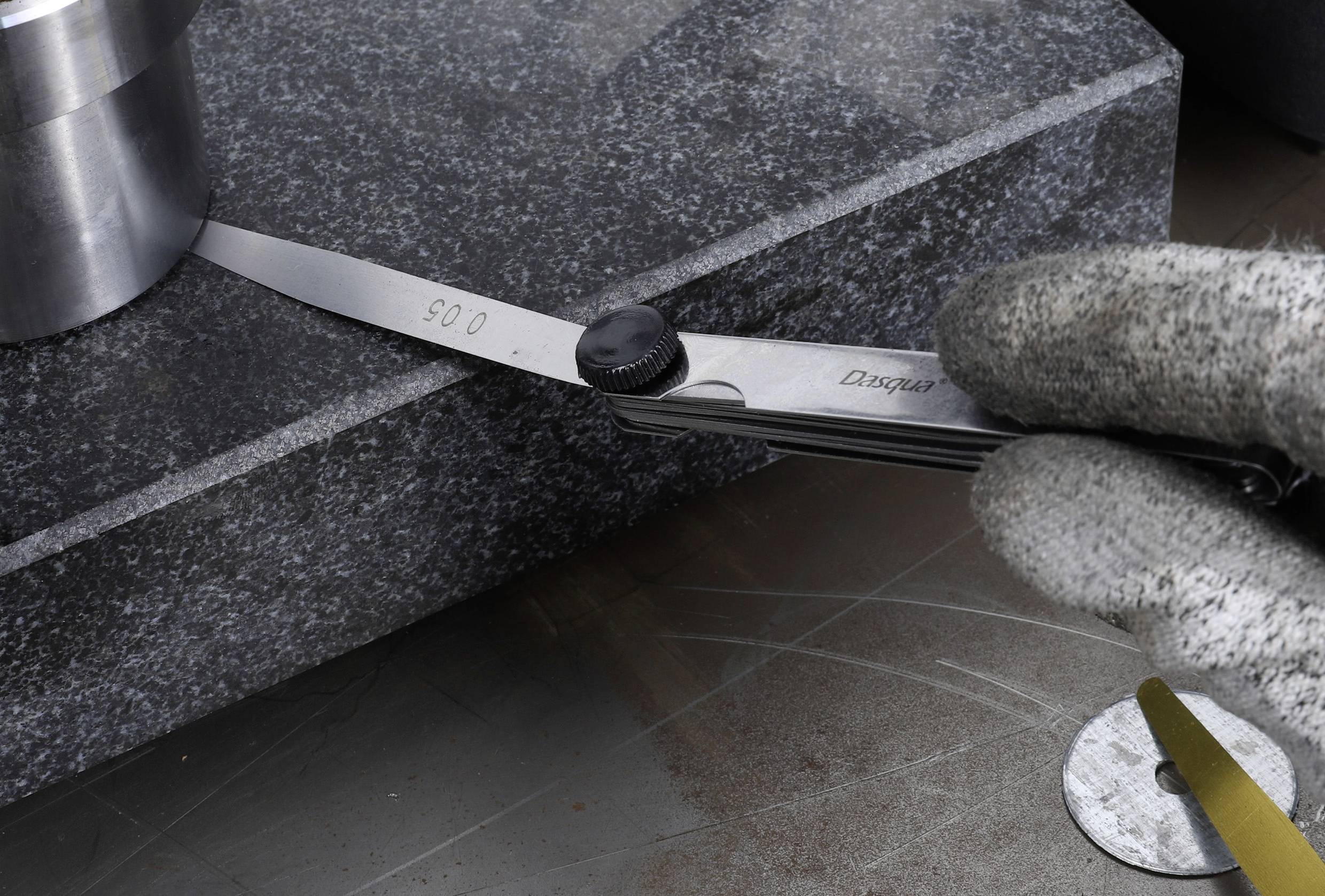 Ręka w rękawicy pomiarowej używa czujnika zegarowego do pomiaru odległości między metalowym walcem a płytą granitową.