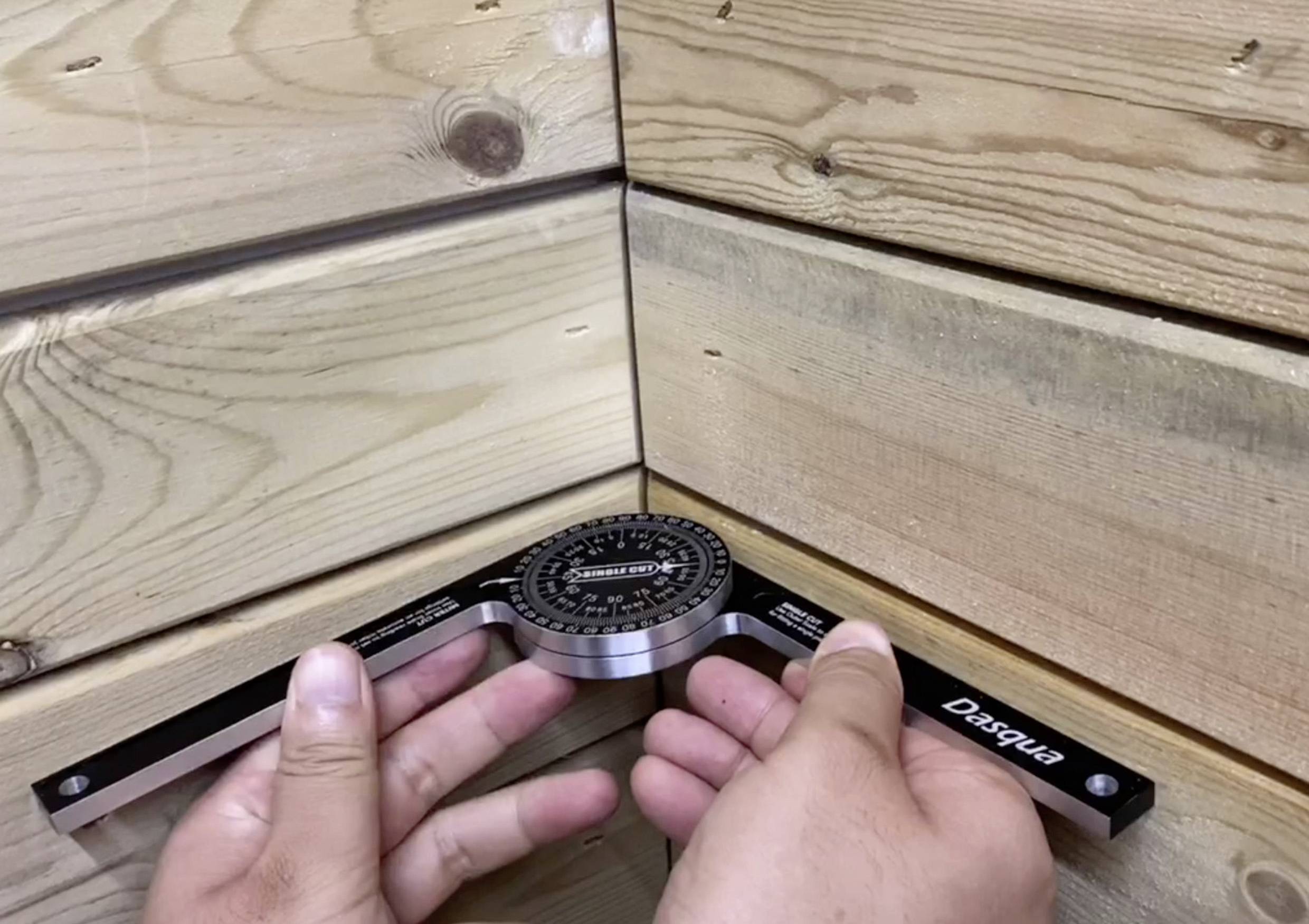 Dwie ręce trzymają kątomierz w narożniku dwóch drewnianych ścian, aby zmierzyć kąt desek.