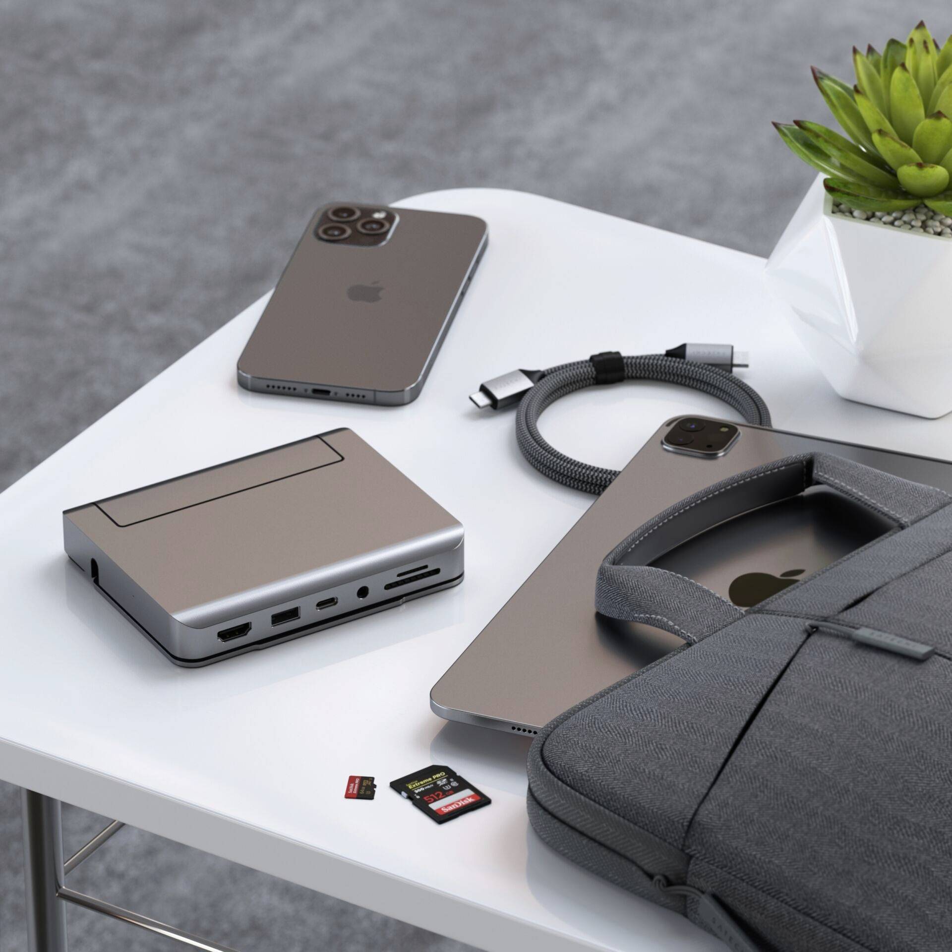 Na stole leżą smartfon, laptop, zegarek, elektroniczne urządzenie i karta pamięci obok małej doniczki z rośliną.