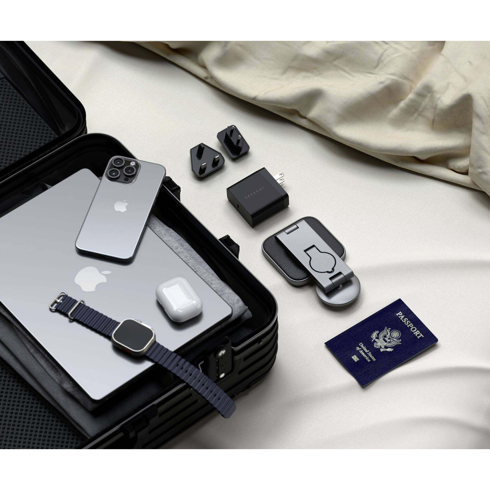 Walizka podróżna z elektroniką: smartfon, laptop, kable ładujące, adapter, smartwatch, słuchawki douszne i paszport ułożone na łóżku.