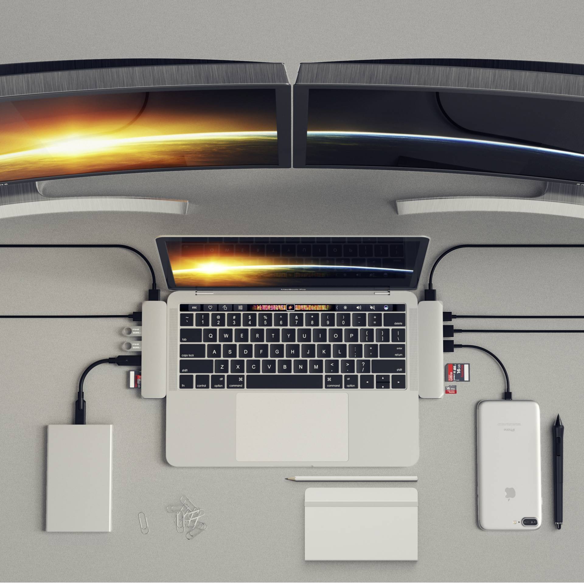 Stanowisko pracy z laptopem podłączonym do dwóch dużych zakrzywionych monitorów. Na biurku leży smartfon, zewnętrzny dysk twardy oraz notes.