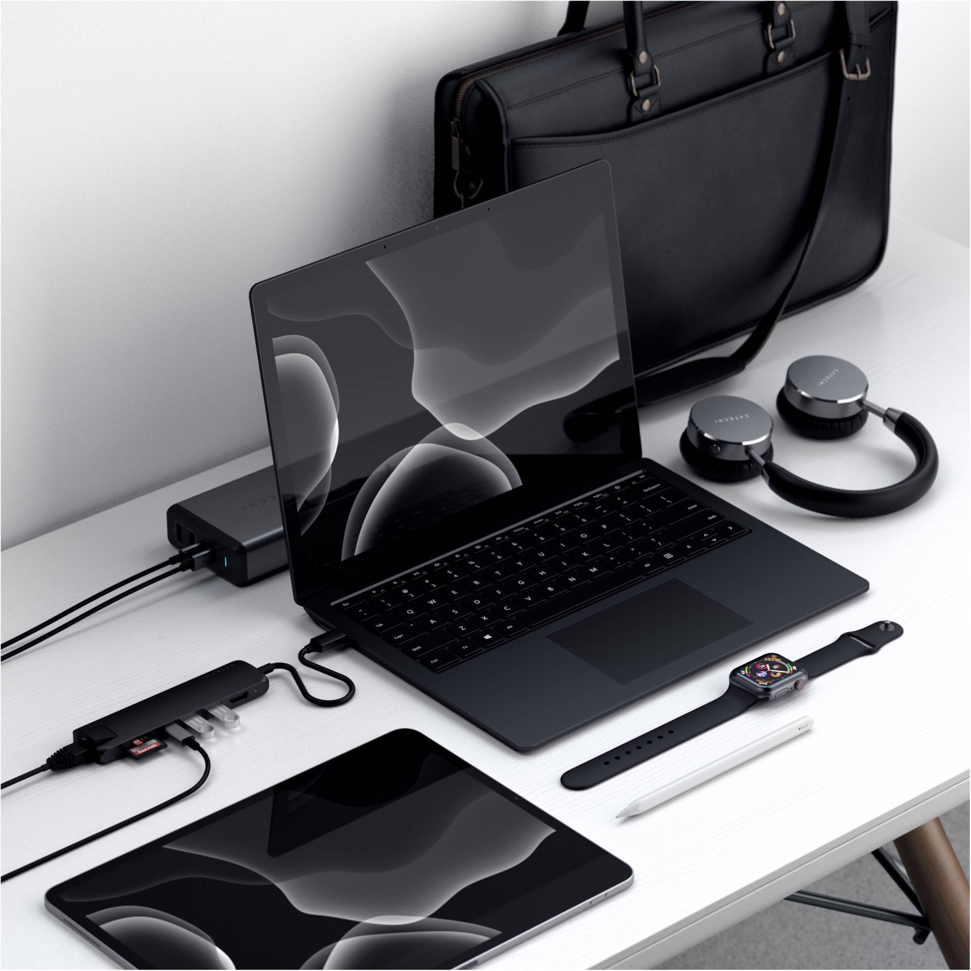 Kilka czarnych urządzeń elektronicznych na stole: laptop, tablet, smartwatch, słuchawki; obok czarna torba.