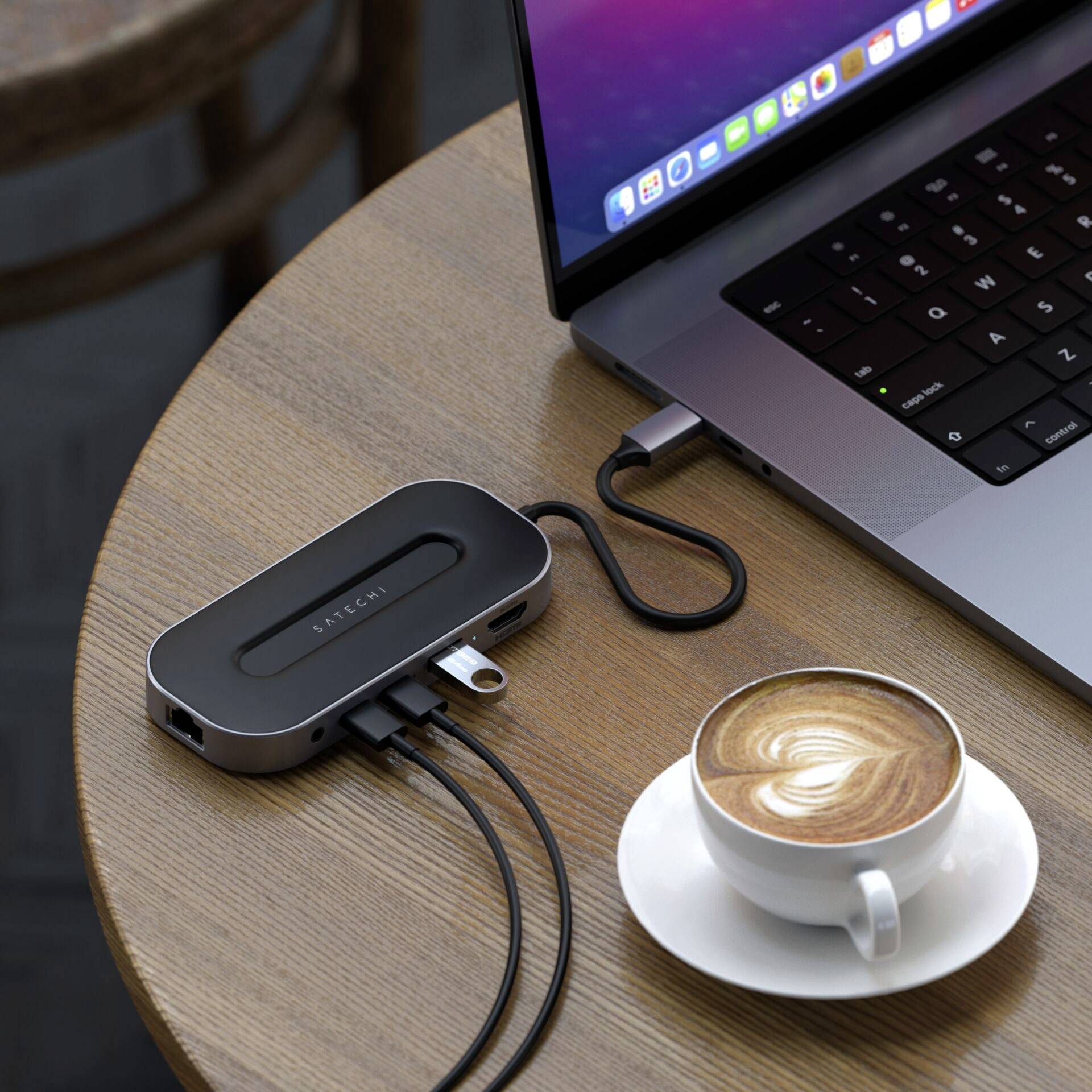 Laptop jest umieszczony na drewnianym stole obok filiżanki cappuccino oraz czarnego rozgałęziacza USB-C z podłączonymi kablami.