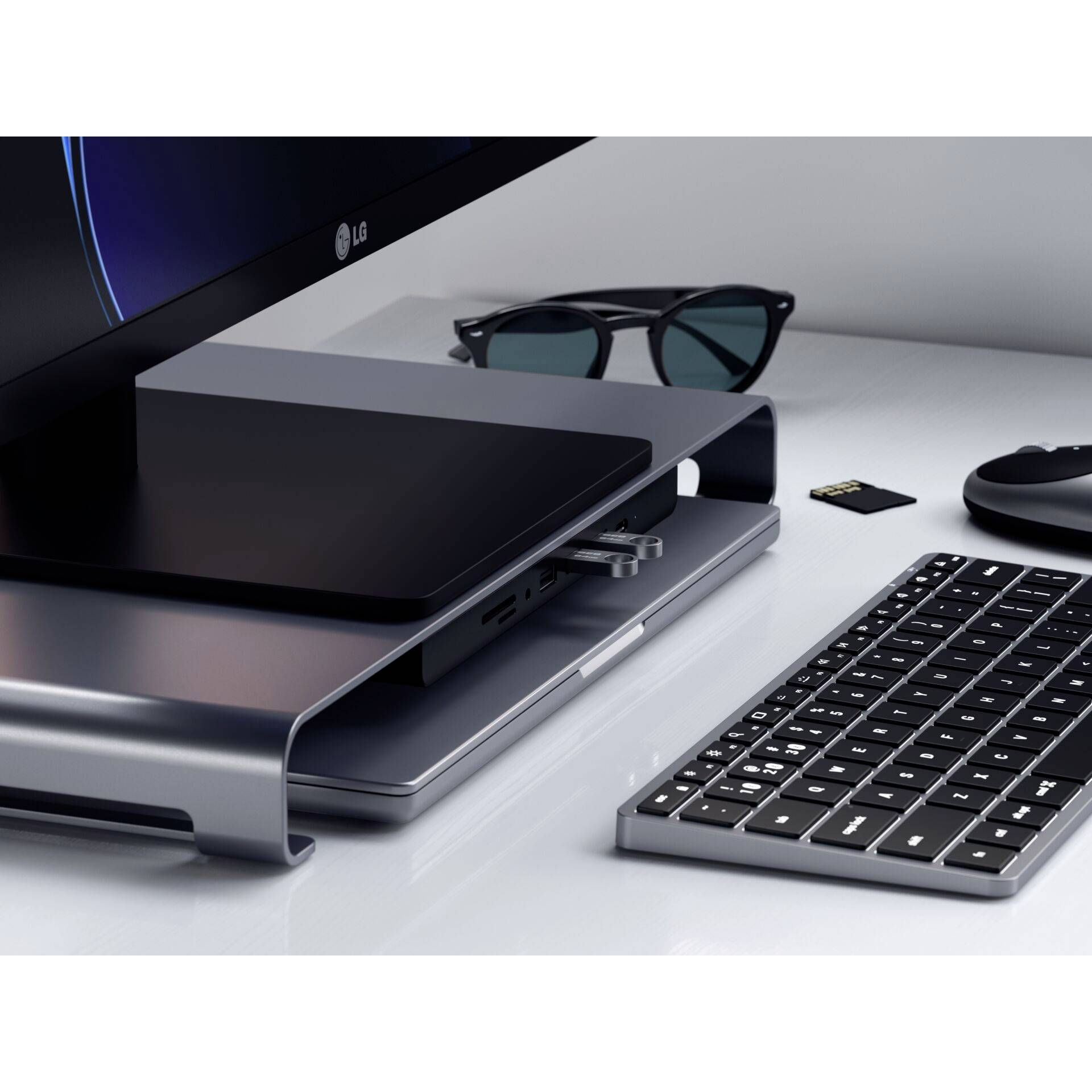 Biurko na stojąco z laptopem, na którym umieszczony jest stojak na laptopa, obok leżą okulary, pendrive, klawiatura i mysz, wszystko na białym biurku.