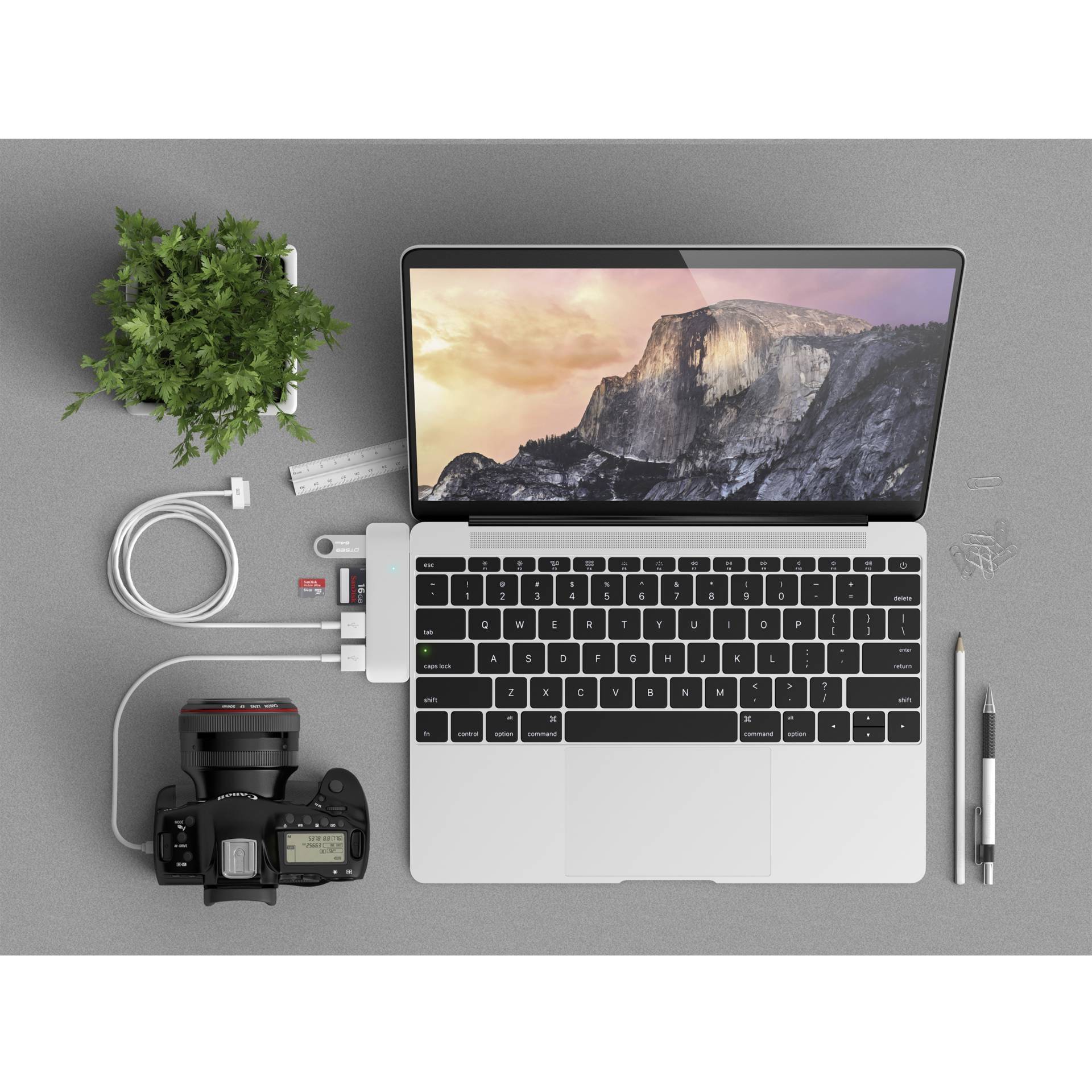 Laptop z tapetą przedstawiającą góry, otoczony kamerą, rośliną, kablami USB i długopisem na biurku.