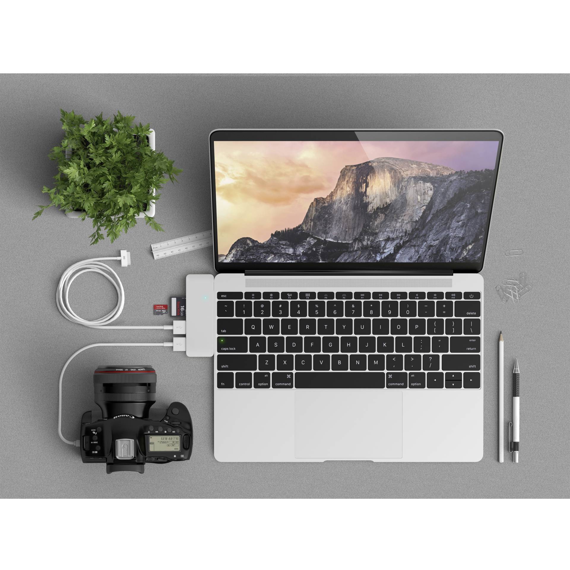 Laptop na biurku z podłączoną kamerą, smartfonem, długopisem, słuchawkami i rośliną; ilustruje środowisko pracy.