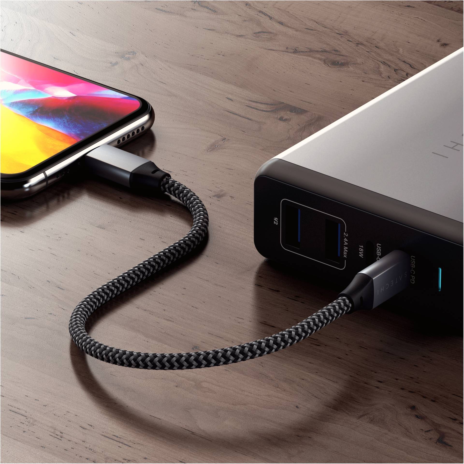 Smartfon jest podłączony plecionym kablem do powerbanku, który leży na drewnianym stole.