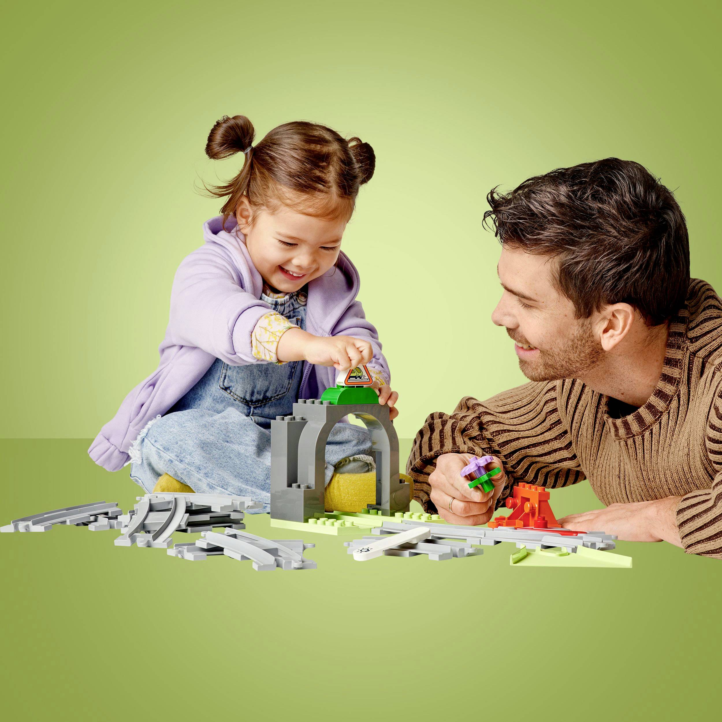 Dziecko i dorosły wspólnie budują zabawkową konstrukcję z klocków na stole, na zielonkawym tle.