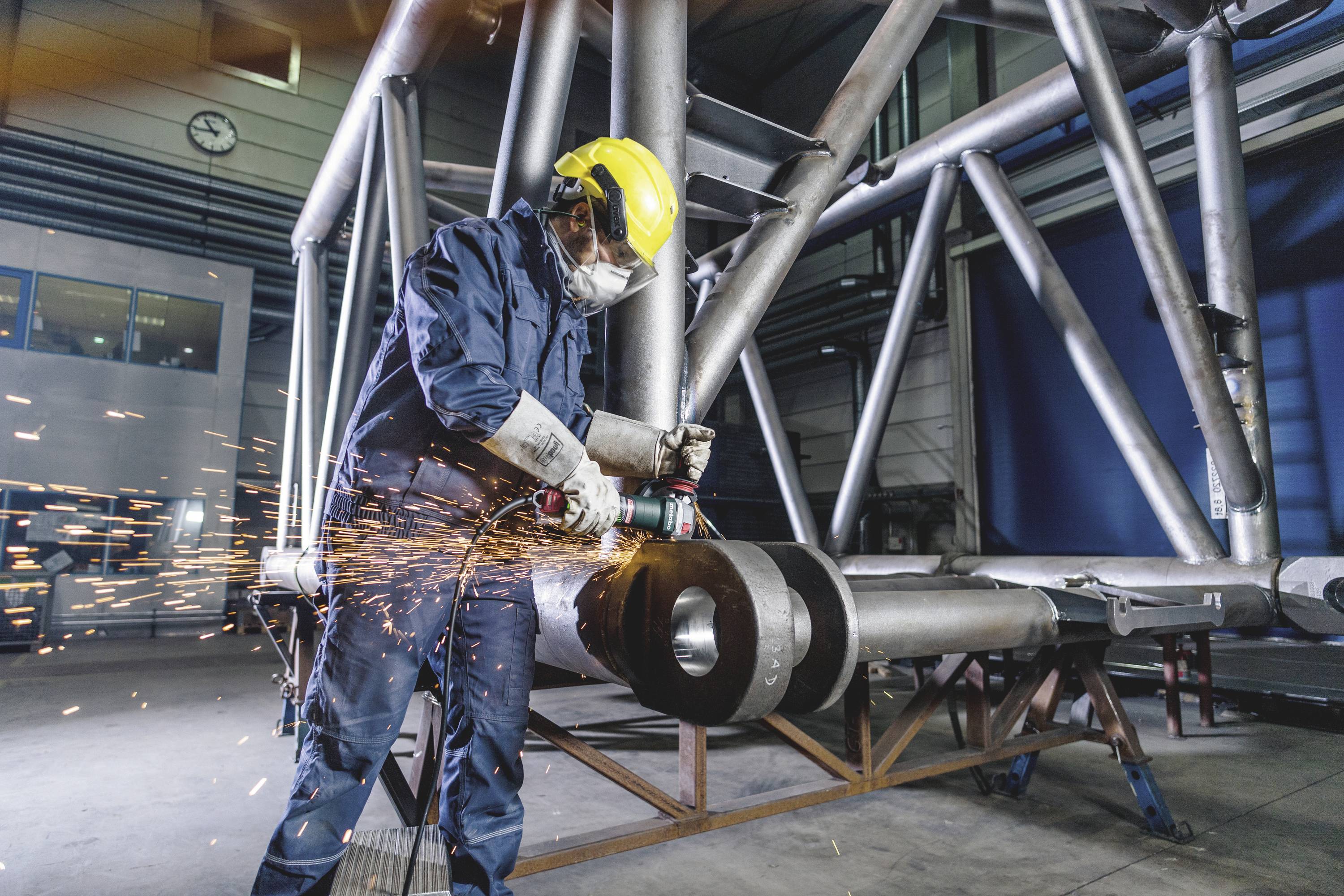 Robotnik w odzieży ochronnej szlifuje metal w hali fabrycznej, a wokół niego latają iskry. W tle widoczne są stalowe belki.