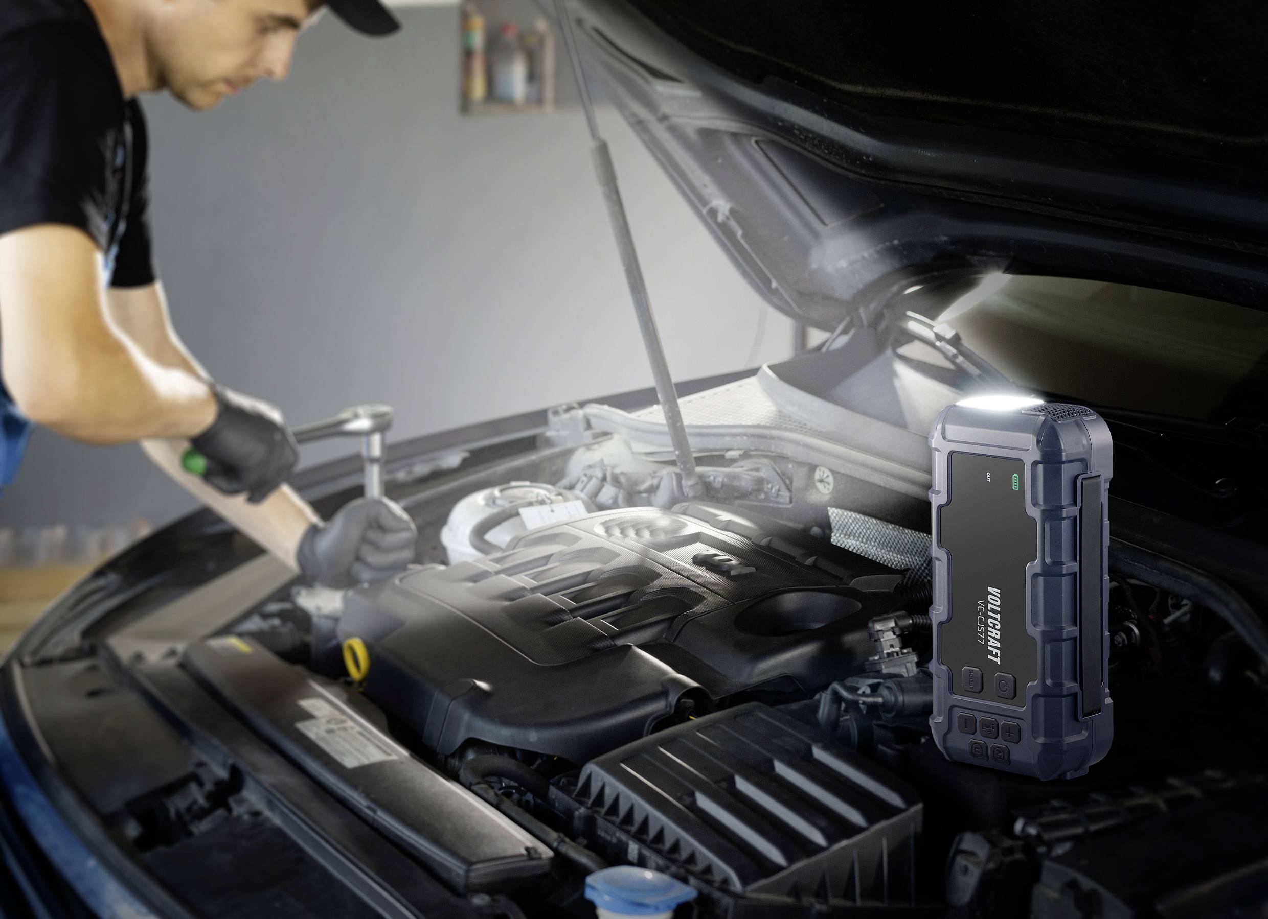 Mechanik naprawia silnik samochodu przy otwartej pokrywie silnika. Na pierwszym planie stoi przenośny rozrusznik.