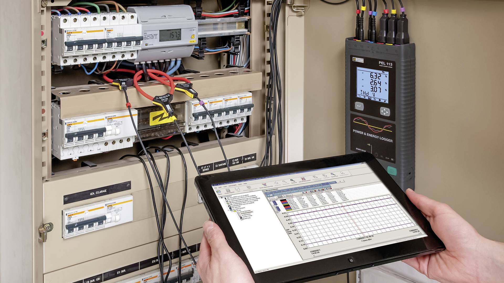 Osoba trzyma tablet przed otwartą rozdzielnicą elektryczną. Tablet wyświetla dane, podczas gdy przyrząd pomiarowy po prawej stronie wskazuje wartości elektryczne.