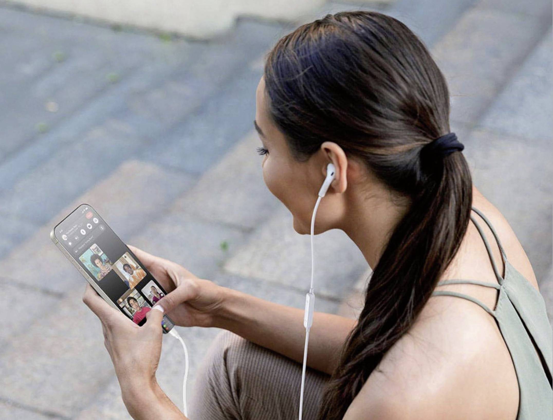 Kobieta siedzi na zewnątrz i prowadzi wideorozmowę na cztery osoby za pomocą smartfona. Ma słuchawki douszne i uśmiecha się.