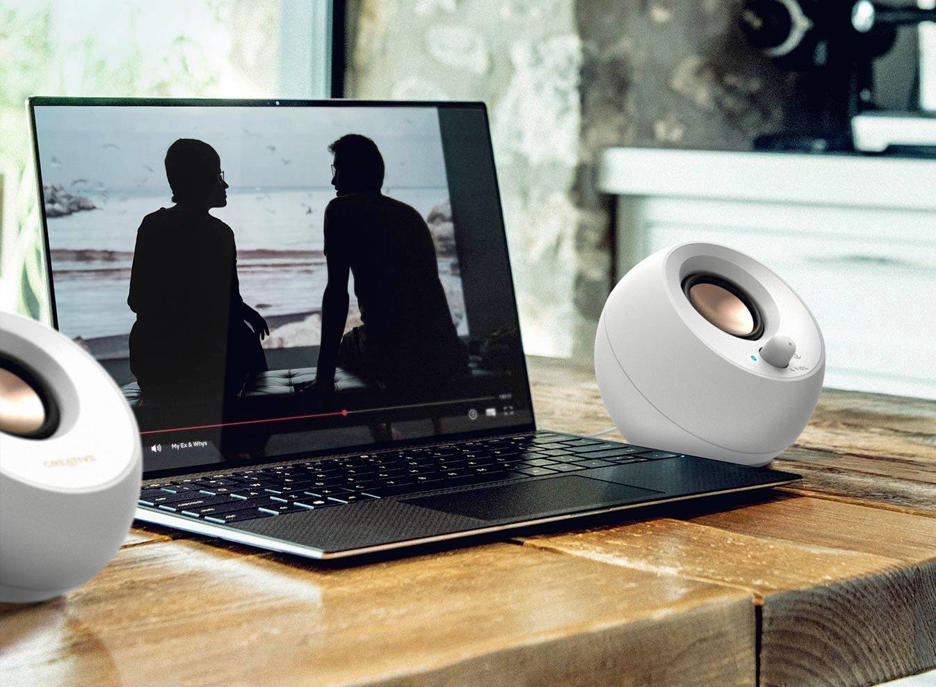 Laptop z filmem przedstawiającym dwie osoby na plaży, otoczony dwoma białymi głośnikami, stoi na drewnianym stole.