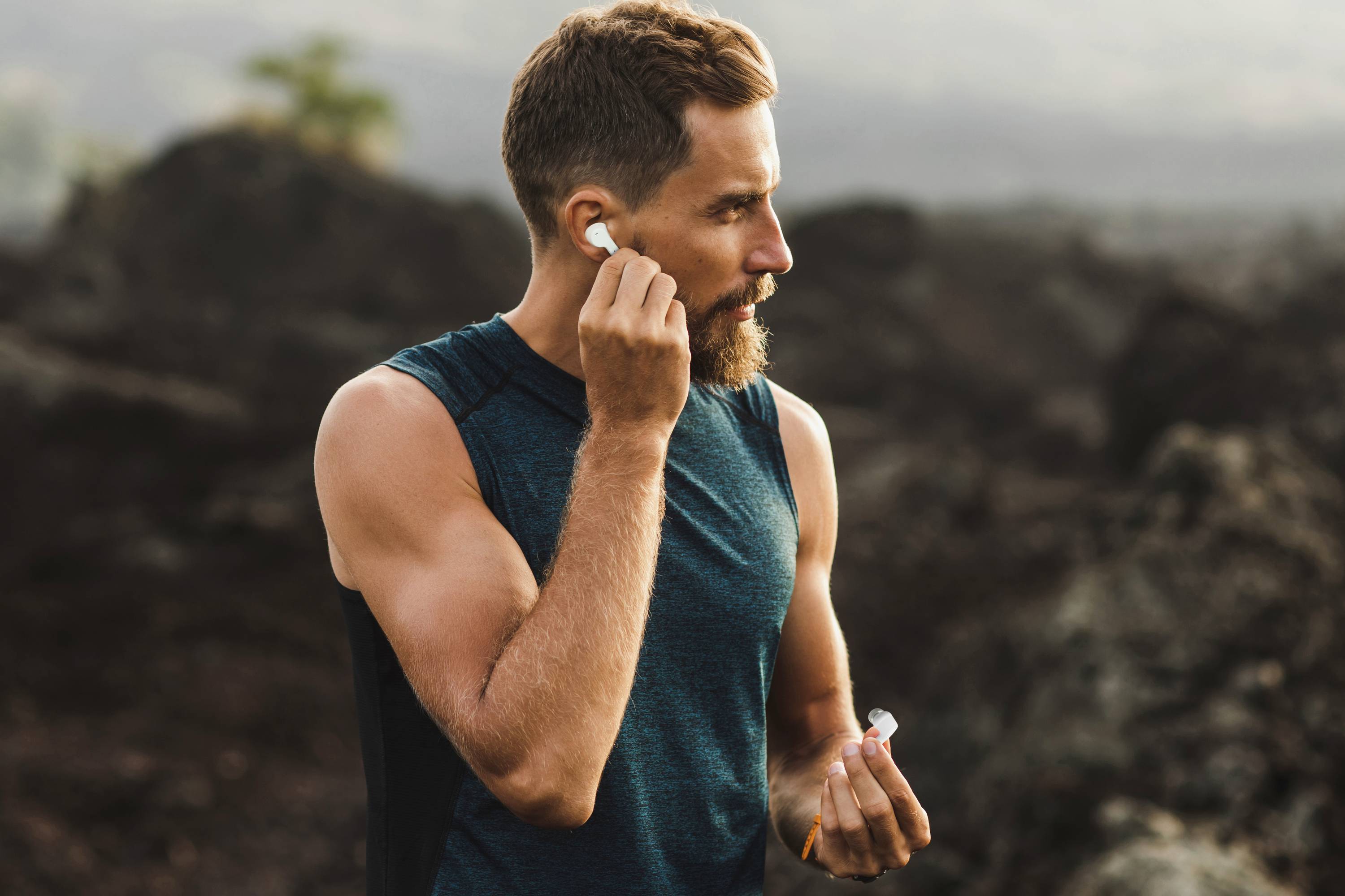 Mężczyzna w bezrękawnej koszulce sportowej wkłada bezprzewodowe słuchawki douszne, stojąc na zewnątrz w skalistym, naturalnym otoczeniu.