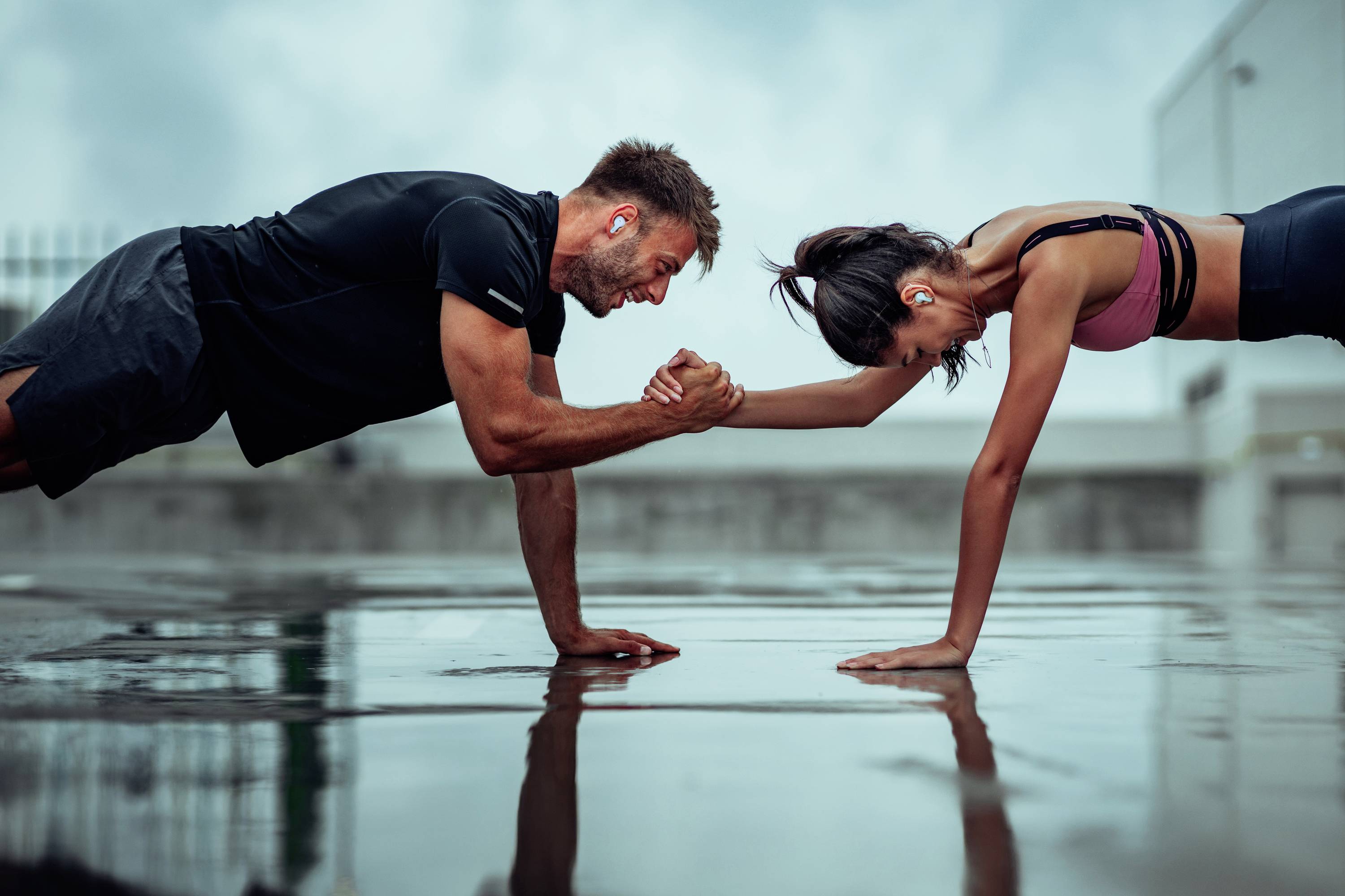 Mężczyzna i kobieta razem wykonują deskę (plank) na mokrej powierzchni. Oboje są ubrани w strój sportowy i mają zatyczki do uszu.
