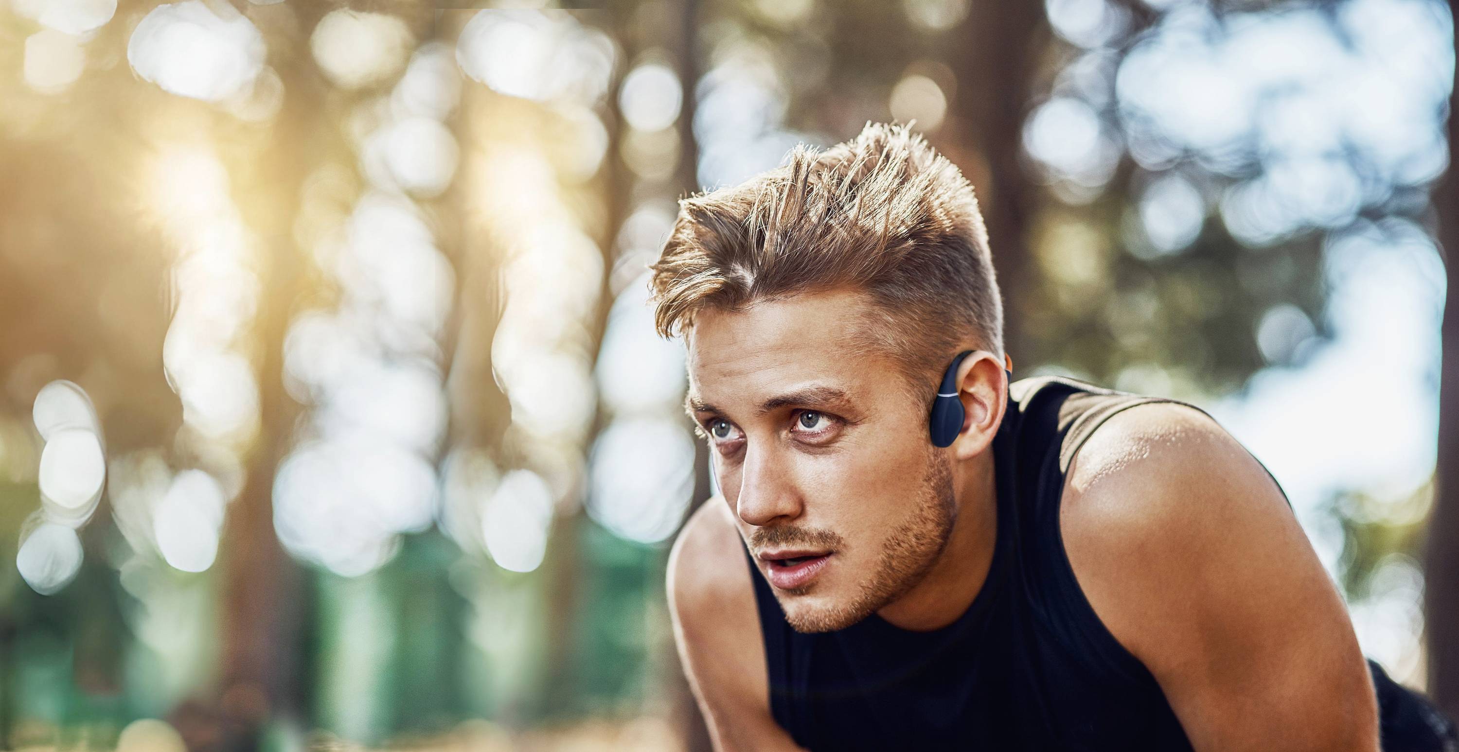 Mężczyzna w stroju sportowym i słuchawkach patrzy skoncentrowany w dal podczas treningu na świeżym powietrzu. W tle widoczne są drzewa.