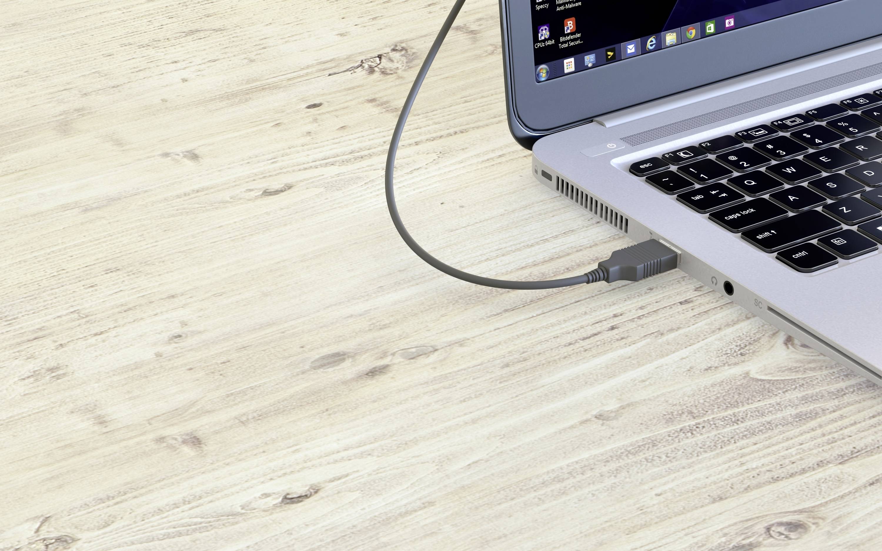 Laptop na drewnianym stole z podłączonym kablem USB. Ekran jest częściowo widoczny i wyświetla otwarte programy.