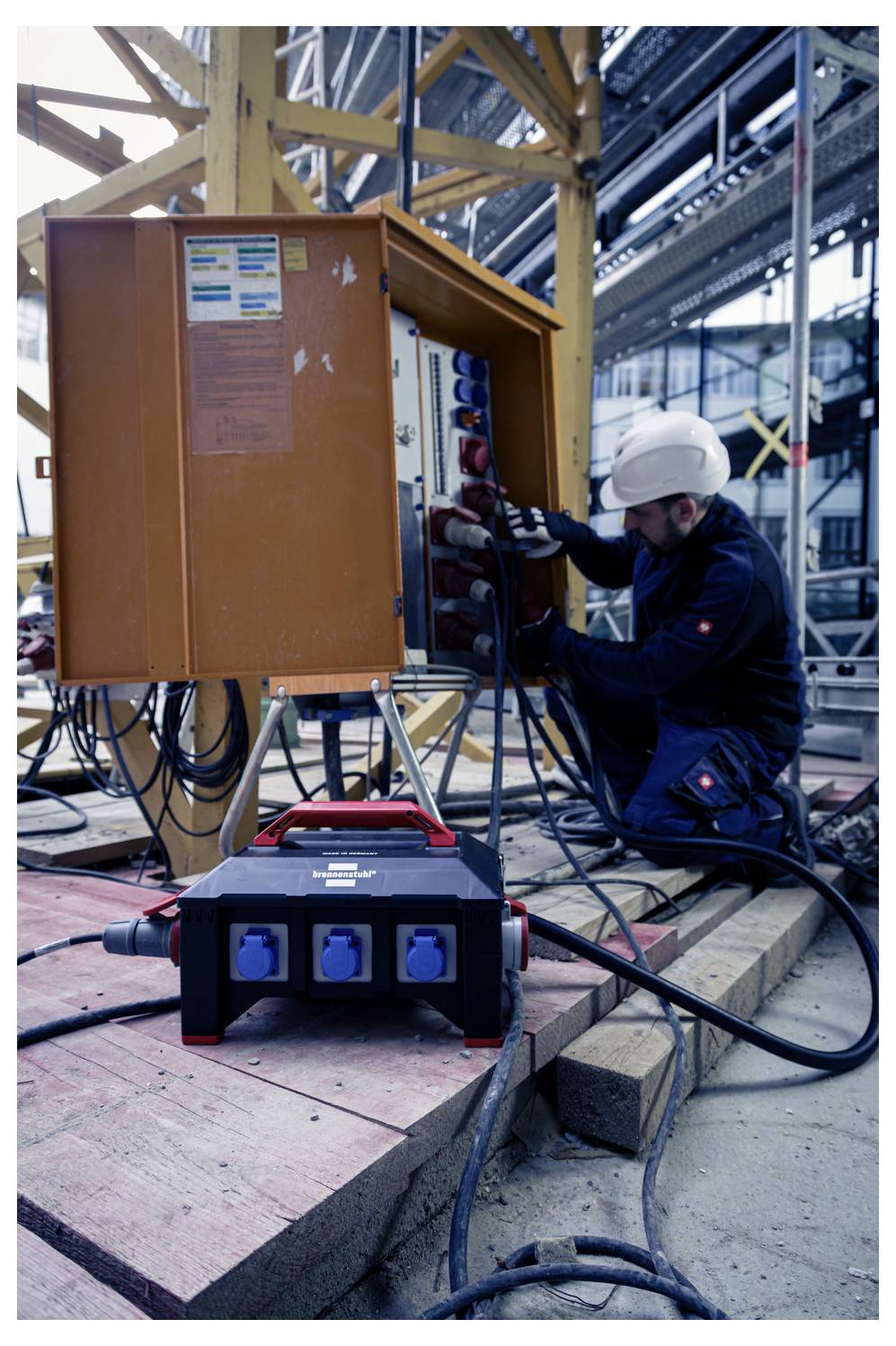 Pracownik budowlany w kasku wdrażający okablowanie szafki sterowniczej elektrycznej na terenie obiektu przemysłowego, z przenośnym skrzyniem rozdzielczym zasilającym na pierwszym planie.