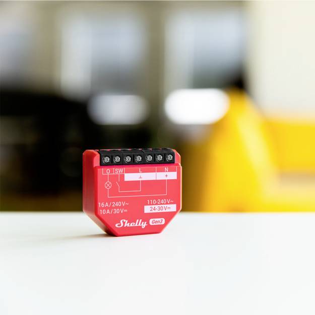 Czerwone przekaźnikowe gniazdo Shelly z zaciskami przyłączeniowymi. Oznaczone jako '16A/240V'. Na rozmytym tle żółte krzesło.