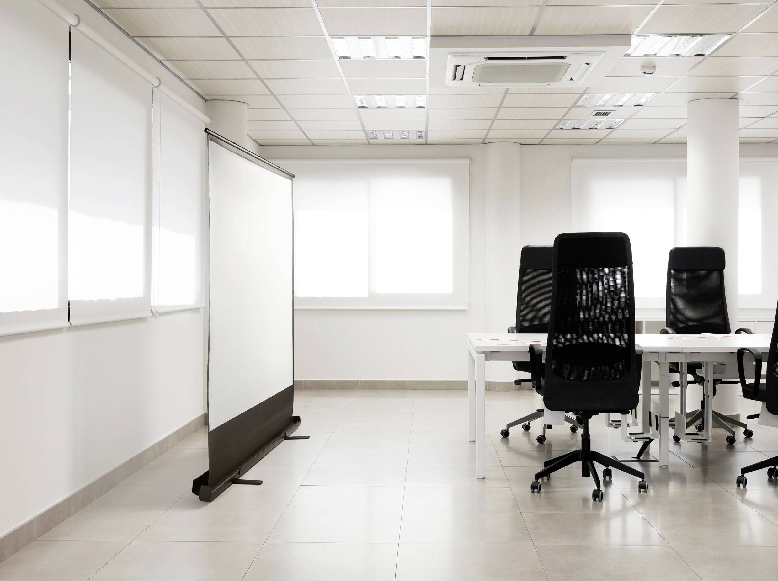 Jasna przestrzeń biurowa z pustym ekranem projektora po lewej stronie oraz trzema czarnymi krzesłami biurowymi przy białych biurkach po prawej stronie.