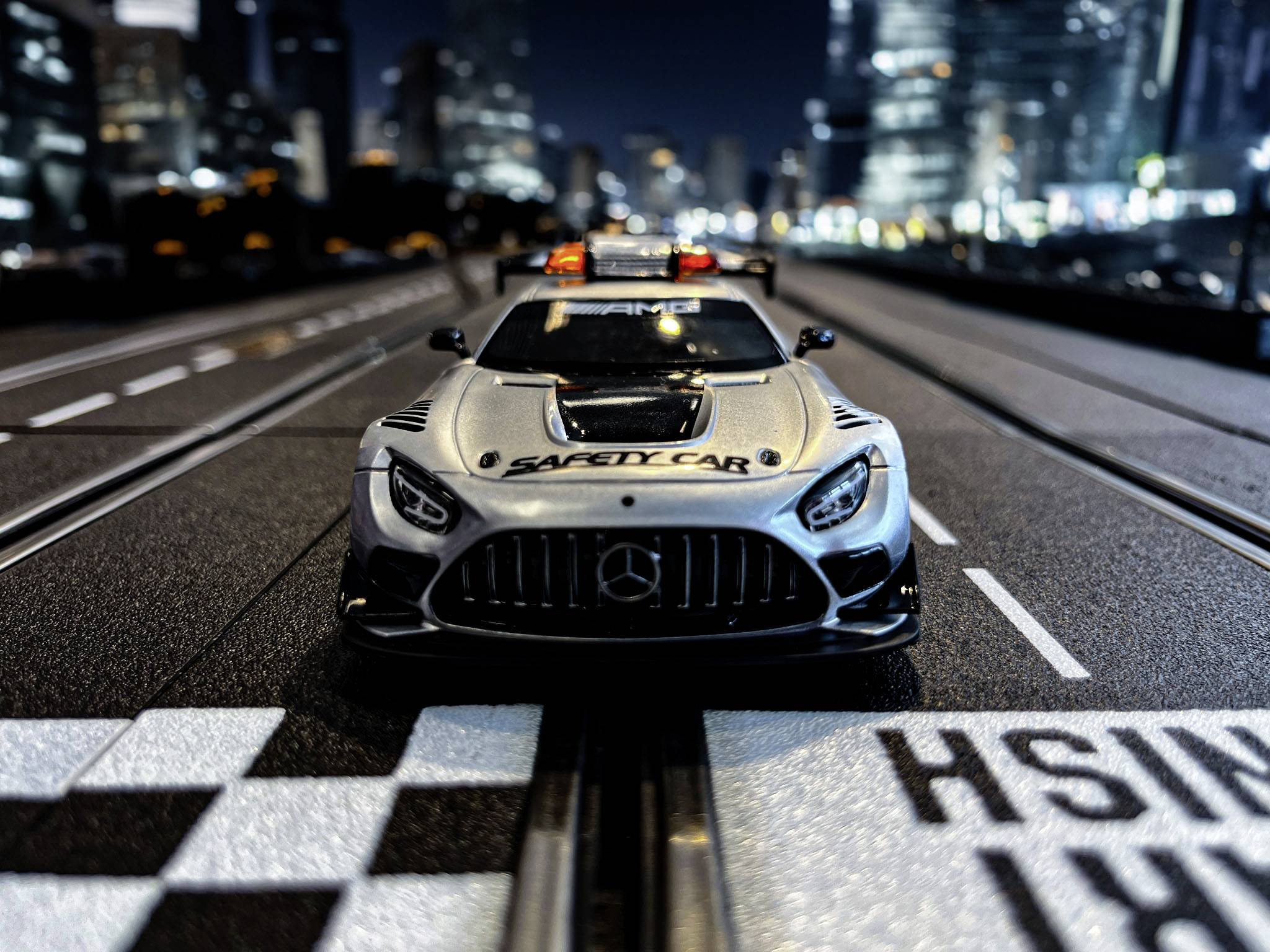 Miniaturowy samochód wyścigowy z napisem 'Safety Car' jedzie po oświetlonej trasie wyścigowej na tle rozmytego krajobrazu miejskiego.