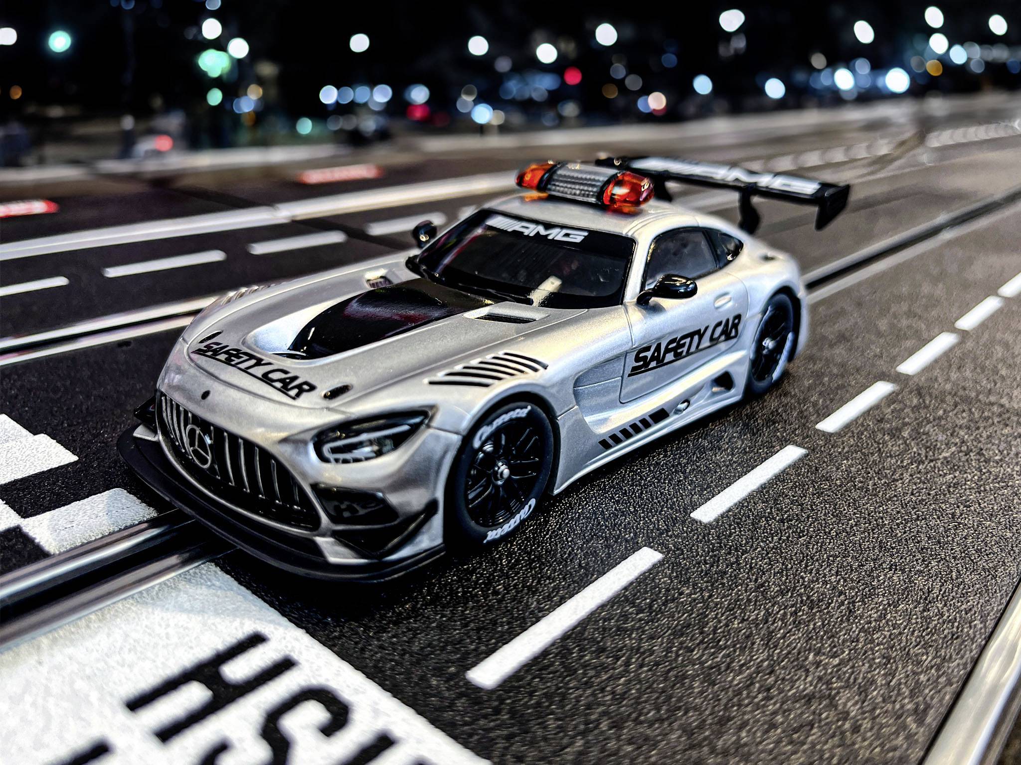 Srebrny samochodzik zabawkowy z napisem 'Safety Car' stoi na torze wyścigowym nocą. W tle widoczne są podświetlone budynki.