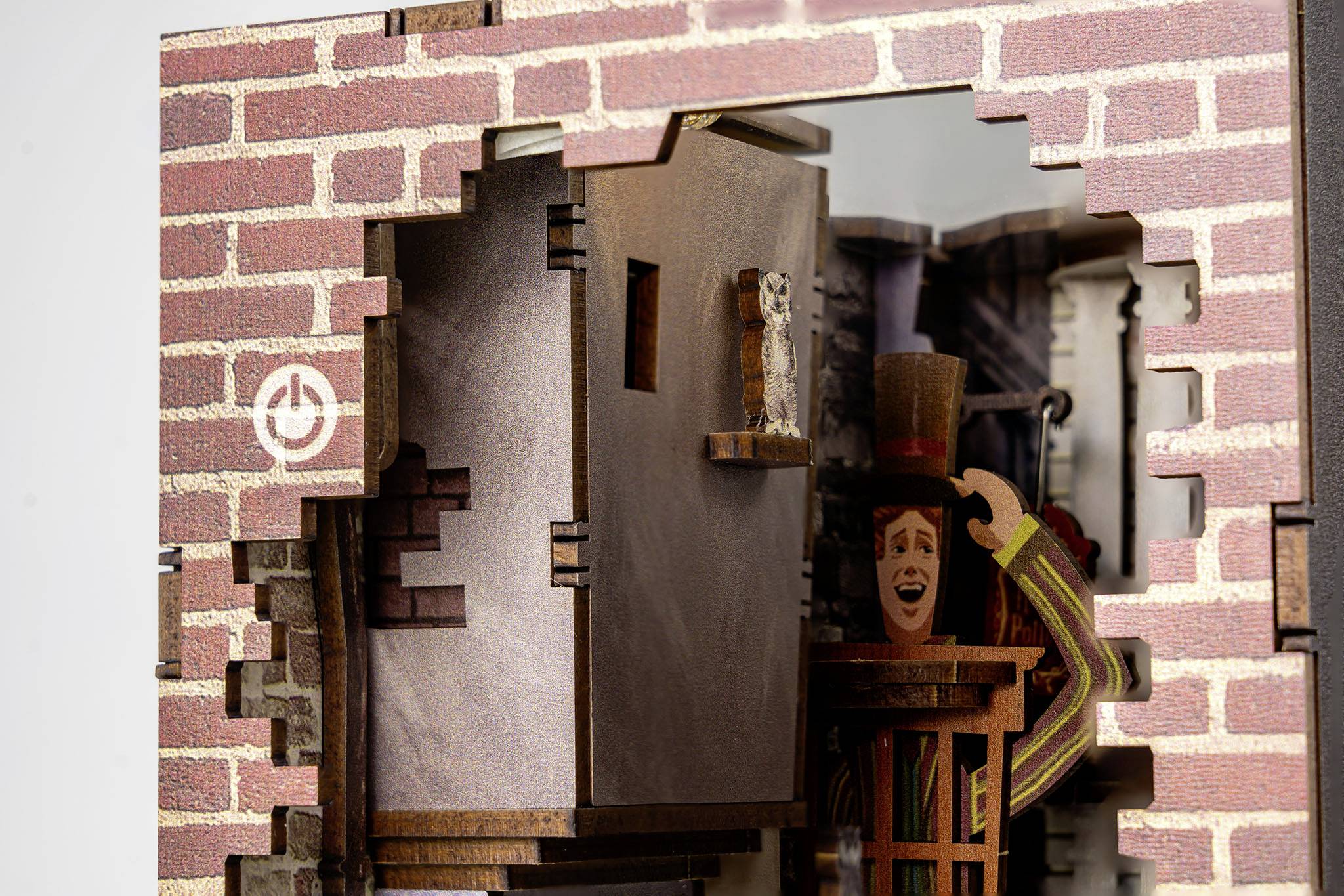 Model domu z odkrytą ścianą z cegły. Figuratywny mężczyzna w prążkowanym garniturze podnosi śmiejąc się kapelusz. Małe zwierzę na gzymsie.