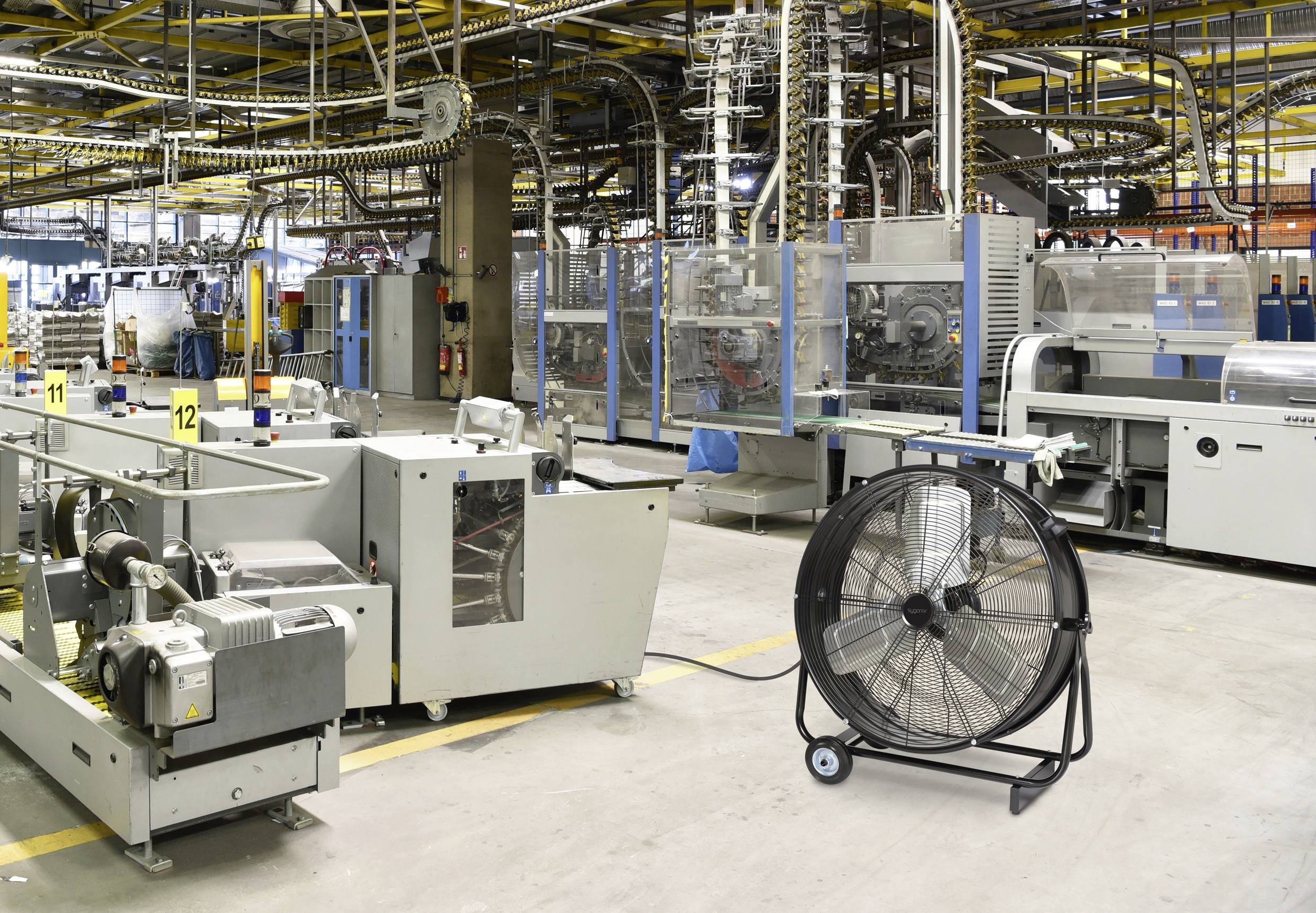 Zakład przemysłowy z różnymi maszynami, w tym duży wentylator w pierwszym planie, stojący na wózku jezdnym.