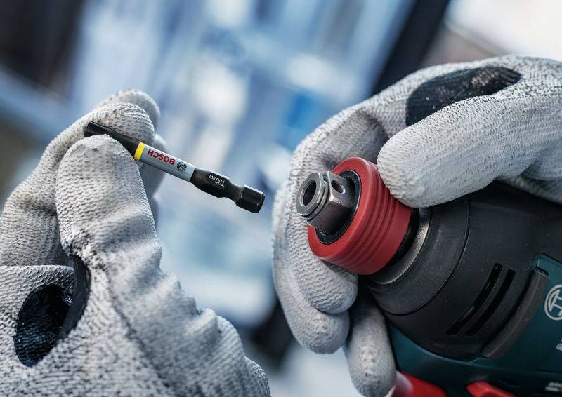 Osoba w szarych rękawiczkach wkłada bit do akumulatorowego wkrętaka. Bit wskazuje markę 'Bosch'.