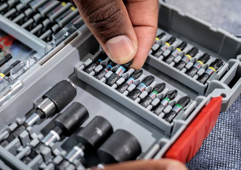 Ręka wybiera wiertło z zestawu w plastikowym pudełku. Pudełko zawiera liczne wiertła i końcówki, które są starannie posortowane.