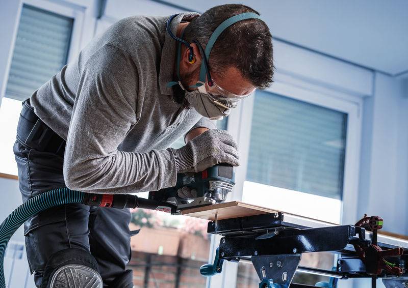 Mężczyzna w odzieży ochronnej pracuje w warsztacie, używając elektrycznej frezarki do obrabiania kawałka drewna na zamontowanym stole.