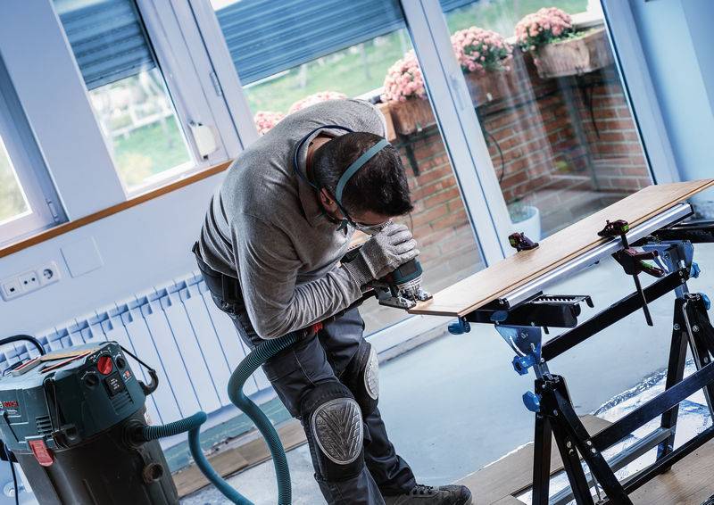 Osoba pracowicie koncentruje się na desce drewnianej, używając elektronarzędzia w warsztacie.