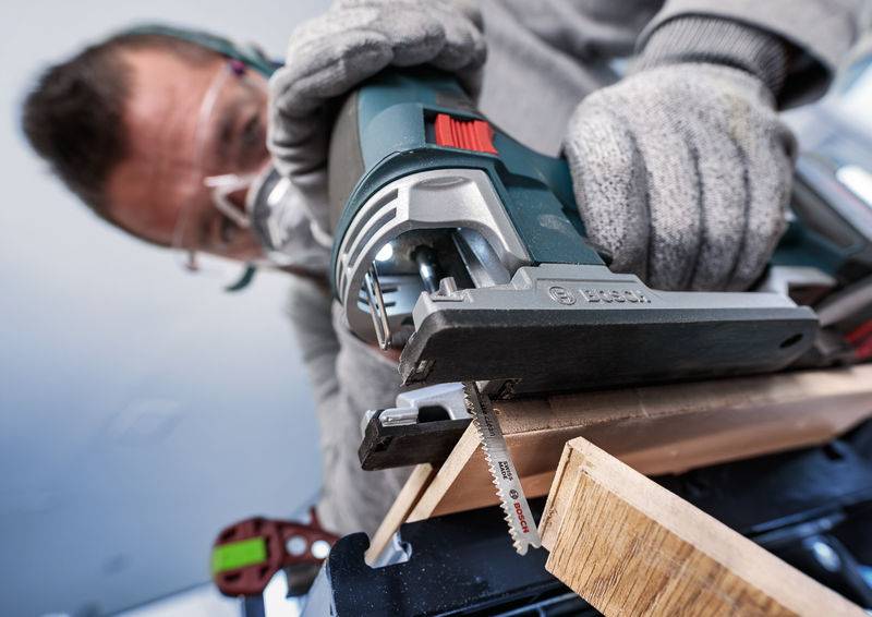 Rzemieślnik przecina kawałek drewna elektryczną wyrzynarką. Nosi rękawice ochronne i okulary ochronne. Fokus jest na pracy piłowania.