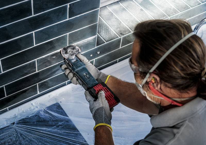 Osoba precyzyjnie ucina czarne płytki za pomocą elektronarzędzia. Podczas pracy nosi okulary ochronne i rękawice.