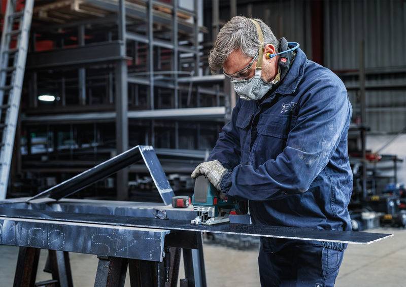 Mężczyzna w stroju roboczym szlifuje metal elektronarzędziem w warsztacie. Nosi odzież ochronną, w tym maskę i ochronniki słuchu.