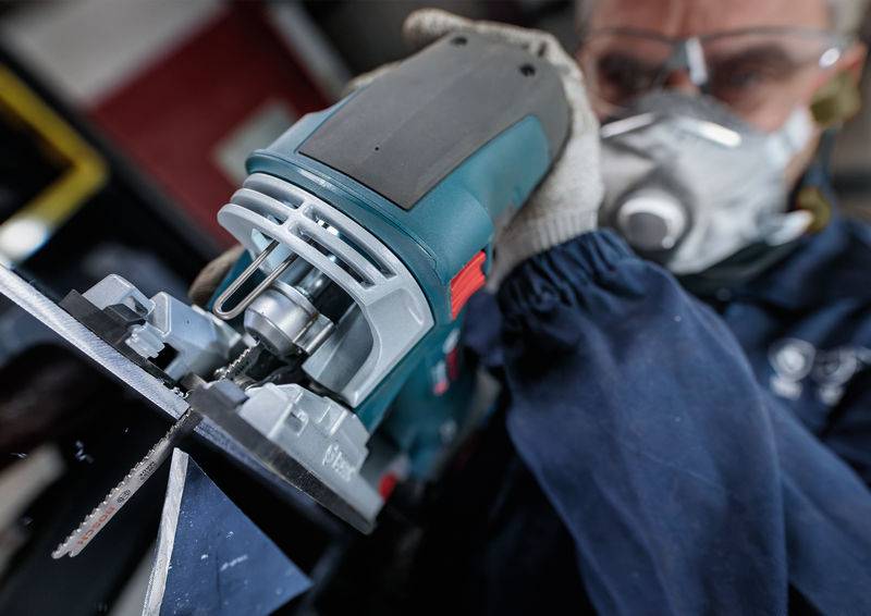 Osoba w okularach ochronnych i masce na twarz używa piły elektrycznej do cięcia metalu. W tle widoczny jest warsztat i otoczenie pracy.