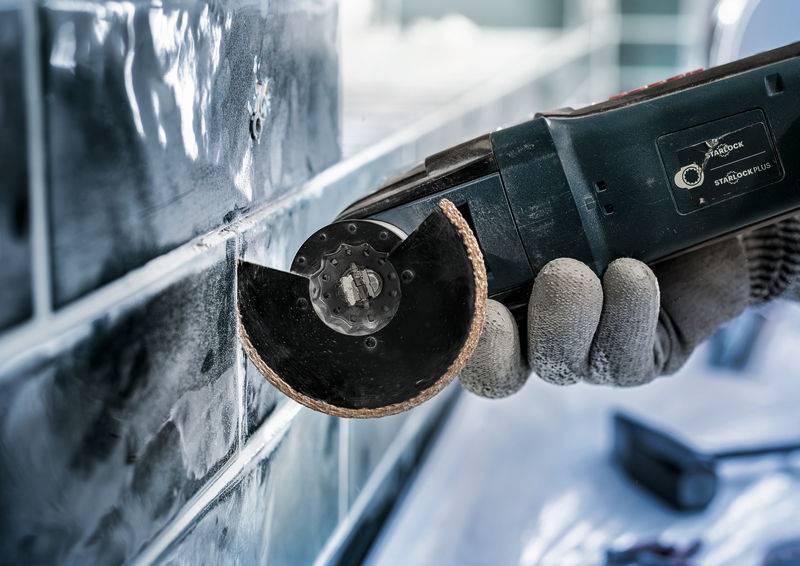 Ręka trzyma obracającą się maszynę tnącą, która obrabia spoiny między błyszczącymi płytkami ściennymi.