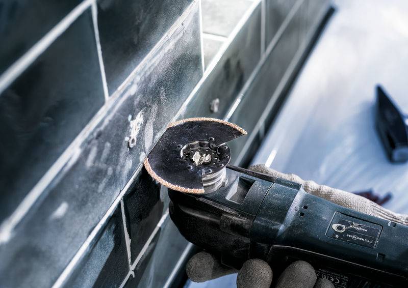 Osoba pracuje z szlifierką kątową, aby ciąć płytki na ścianie. Rękawice chronią dłonie podczas pracy.