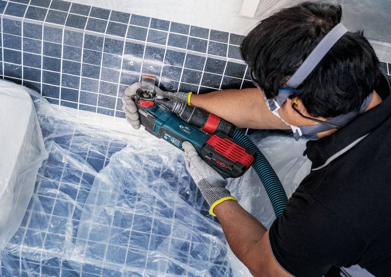 Osoba wiertli elektrycznym narzędziem w kafelkowaną ścianę. Ubranie ochronne i zabezpieczenie przed pyłem są stosowane.