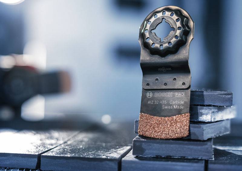 Oscylujące ostrze piły spoczywa ukośnie na ułożonych w stos płytkach. Na ostrzu widnieje napis „Carbide Swiss Made".