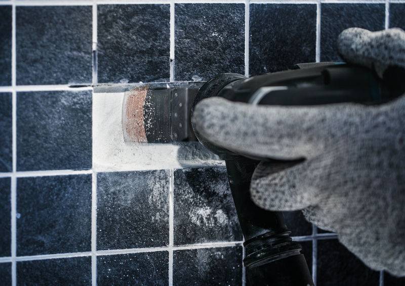 Dłoń w szarym rękawiczce używa elektronarzędzia do usuwania fugi między ciemnymi płytkami.