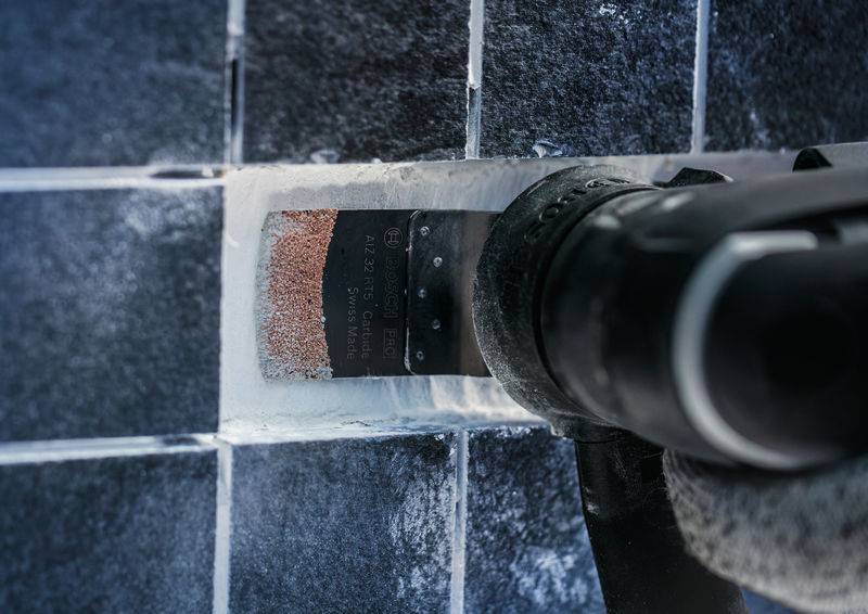 Narzędzie usuwa fugi z ciemnej ściany kafelkowej. Tarcza tnąca usuwa białą zaprawę fugową.