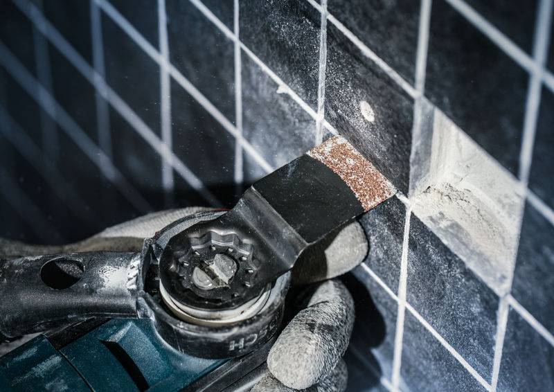 Dłoń w rękawicy trzyma oscylujące narzędzie, które obrabia spoinę między ciemnoszarymi płytkami. Unosi się pył.