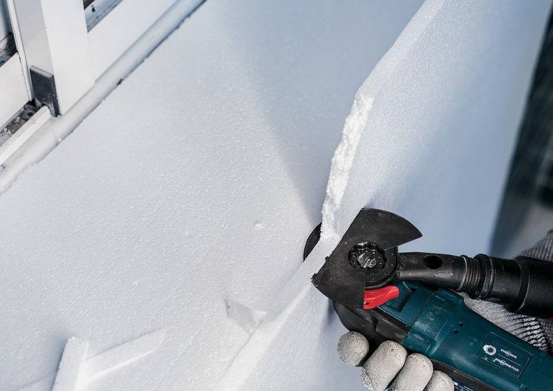 Rzemieślnik ucina grubą płytę styropianową kątówką przy ścianie. Akcent położony jest na precyzyjnym wykonaniu pracy.