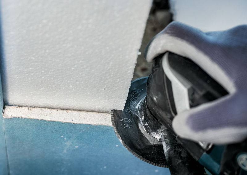 Dłoń w rękawicy trzyma narzędzie elektryczne, które przecina styropian. Zbliżenie ukazuje precyzyjną pracę przy krawędzi ściany.